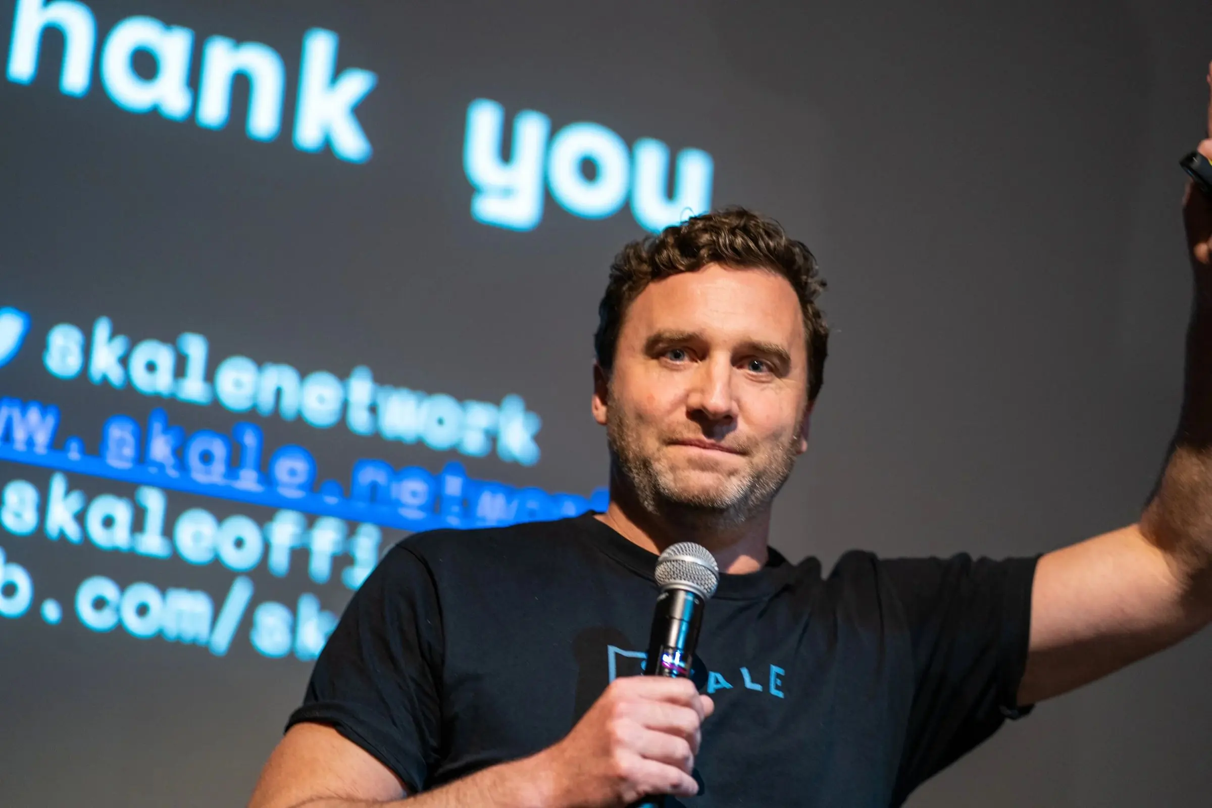 A speaker with a microphone raising his arm in front of a projected thank-you slide
