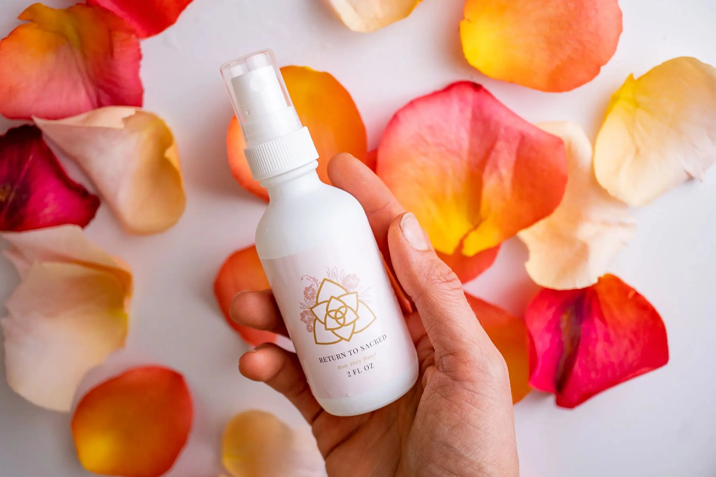 A hand holding a white serum bottle over scattered rose petals